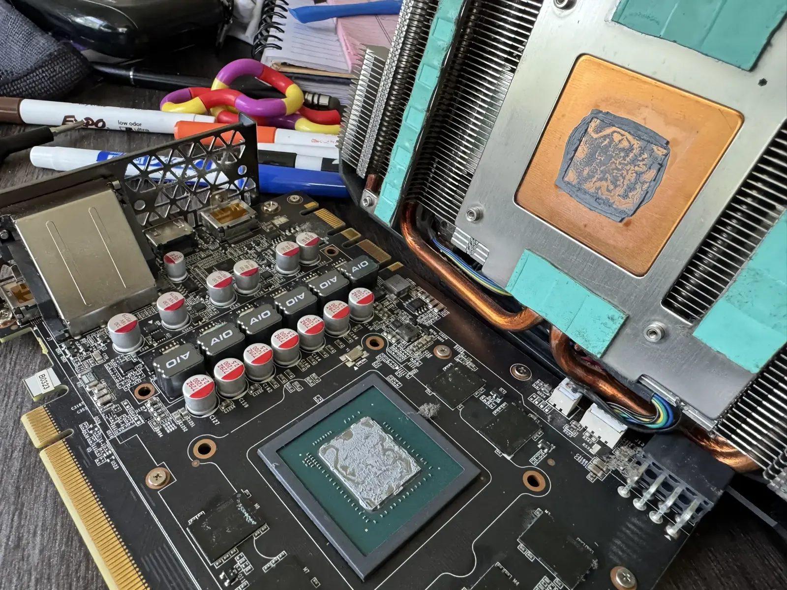 A disassembled graphics card and heatsink on a workbench, with thermal paste visible on the chip and copper cold plate.
