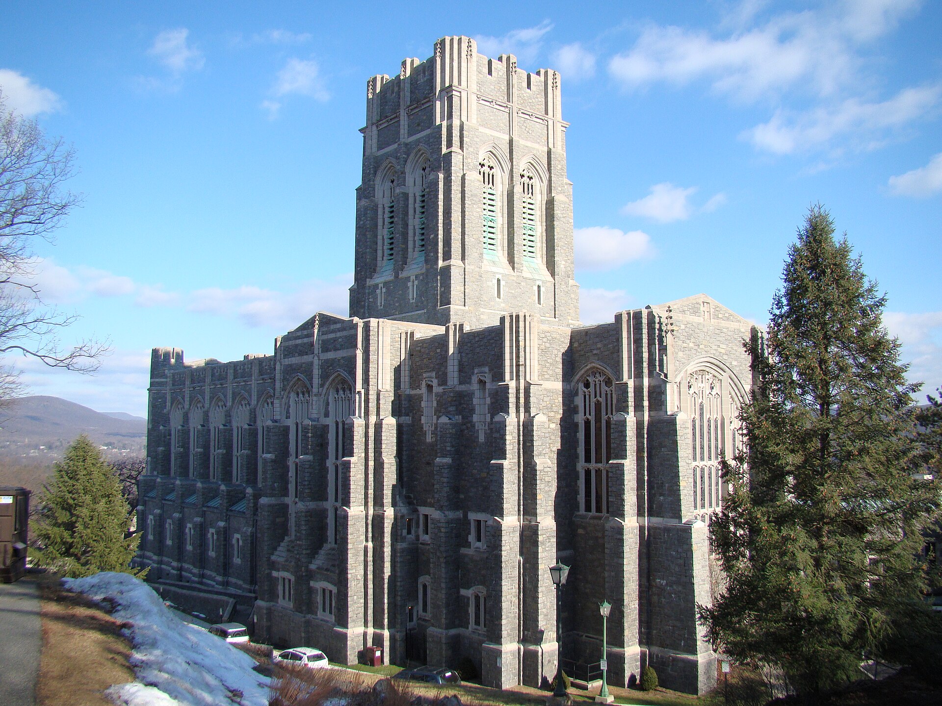 Large gray stone Gothic-style church with a tall square tower, pointed-arch windows, and buttresses, set on a hillside with patches of snow, evergreen trees, and mountains in the background under a bright blue sky with scattered clouds.
